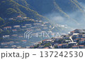 A drone pans over a hillside town nestled between forested mountains. The soft morning mist and golden light create a peaceful, natural ambiance over the rooftops. 137242530