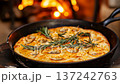 A freshly baked focaccia in a skillet is topped with rosemary and garlic slices, steaming near a wood fire. The cozy setting adds a rustic warmth to the scene. 137242763
