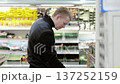 Man in a black jacket inspects groceries in a supermarket's refrigerated section, carefully evaluating fresh produce and comparing choices to fit his needs and budget 137252159