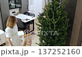 Family members collaborating, positioning vintage christmas tree in modern, sunlit apartment, sharing joyful holiday preparation and strengthening familial bonds 137252160