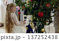 Blond girl in white dress gently adorning christmas tree, carefully placing nutcracker ornament with festive smile, creating warm holiday atmosphere at home 137252438