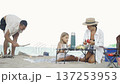 A family spends leisure time at the seaside, having a picnic with fresh fruit and drinks on a sandy beach. The scene captures summer relaxation, bonding, and outdoor fun in a coastal setting 137253953