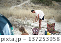 Adult male gathers wood for a campfire on a serene beach, surrounded by camping gear and natural scenery. Peaceful summer day spent enjoying nature and preparing for relaxation 137253958