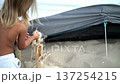 A young child with long blonde hair is seen joyfully playing with sand near a tent on a bright beach day. The image radiates summer vibes, childhood happiness, and natural adventure 137254215