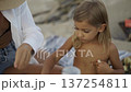 Shirtless child, adult share learning experience on beach, with adult holding knife, child concentrating on task. Scene captures peaceful, natural, educational family interaction 137254811