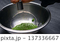 Hand holding a metal grinder above a stainless steel bowl filled with vibrant green spinach mixture, showcasing the grinding process in a kitchen setting 137336667