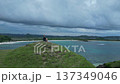 Lady Seated On Cliff Observing Colorful Ocean And Distant Hills 137349046