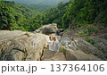 Young woman sitting on rocky trail above lush tropical valley, capturing scenic mountain landscape with smartphone in peaceful outdoor travel setting with wide copy space 137364106
