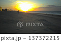 Low-angle wide shot of people walking along a beach during a colorful sunset. Silhouetted figures move peacefully along the shoreline as warm orange and pink hues fill the sky. 137372215