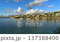 A stunning panoramic view of a tranquil blue bay with elegant white villas nestled on a sunlit hillside under a vast blue sky with soft, drifting white clouds in a Mediterranean setting. High quality 137388400