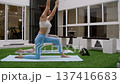 Athletic females follow online class on tablet while changing poses on mats. Minimalist courtyard design creates stylish space for flexibility and strength work. 137416683