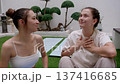 Two young women sit on grass and chat during stretching break in zen courtyard. Hands gesture casually while bonsai trees and lanterns decorate background. 137416685