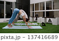 Athletic females stretch spine and shoulders then move into downward pose during guided session. Minimal courtyard space becomes private studio for wellness routine. 137416689