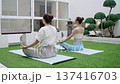 Two fit women practice lotus pose on yoga mats in zen courtyard with bonsai trees. Hands form mudra while stone lantern and plants create peaceful atmosphere. 137416703