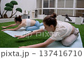 Fit girls perform forward bend stretch on blue mats over green grass surface. Asian style lanterns and bonsai trees in background create calm wellness atmosphere. 137416710