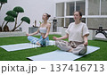 Two young women sit in lotus pose on yoga mats during meditation session in zen courtyard. Hands rest calmly palms up while plants and lanterns surround serene space. 137416713