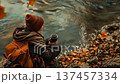 Individual enjoying autumn vibes by the water, sipping coffee amidst colorful fallen leaves 137457334