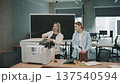 Two people discussing work tasks by a printer in an office setting during a busy workday 137540594