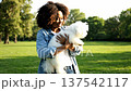 Smiling young afro american woman with curly hair hugging white fluffy maltese dog in sunny green park on blurred trees background. Concept of friendship and pet care 137542117