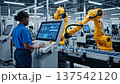 Female afro american engineer in safety glasses operating a digital touch screen control panel near yellow robotic arms on a factory production line. Concept of women in engineering and smart manufact 137542120