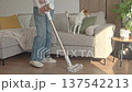 Woman vacuums floor with cordless stick vacuum while dog stands on sofa. Concept of household cleaning 137542213