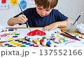 A cute young boy with painted cheeks draws a red heart on white paper, surrounded by paint and brushes. Symbolizes childhood creativity, art, love, and early education. 137552166