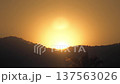 Golden sun disk emerging above silhouetted hills and tree line, casting warm glow across hazy sky at dawn or dusk, serene natural landscape scene 137563026
