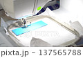 4K Close up of a high-speed computerized embroidery machine stitching a white letter T onto premium blue leather. Precision industrial sewing process and textile manufacturing technology. 137565788