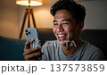 Happy Asian man laughing while using smartphone at home in cozy living room with warm lamp lighting 137573859