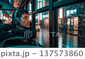 Asian man wearing headphones sitting on public bus looking through rainy window at neon lit city street at night 137573860