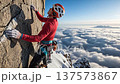 Professional rock climber in red gear ascending mountain cliff face above dramatic cloud layer with alpine peaks in background 137573867