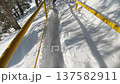 POV walk along a snow-covered metal grating walkway. Deep snow surrounds the path and bright yellow railings on a sunny winter hillside 137582911