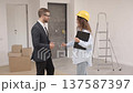 Female designer in helmet with documents giving keys from apartment to young businessman while standing in room 137587397