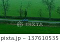 Blue SUV speeding along narrow rural road between green agricultural fields. Fast vehicle moving through countryside farmland landscape bordered by hedgerows and winter trees. Dynamic aerial driving 137610535
