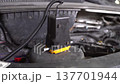 A car part sits on the engine block under the hood of a vehicle. Wires are connected to the part while tools and other components are nearby. This scene shows work in a garage. 137701944