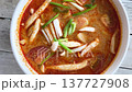 Close-up of a bowl filled with Tom Yam soup featuring mushrooms, green onions, and a flavorful red broth on a rustic wooden surface 137727908