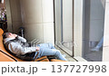 Young man in casual clothing relaxes in a lounge chair at the airport, waiting for his flight, with large windows and contemporary decor in the background 137727998