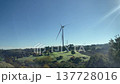 Wind turbines rotate in a bright blue sky, surrounded by lush greenery and open fields, highlighting the integration of renewable energy in nature 137728016