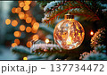 A delicate Christmas ornament hangs from a branch, glowing softly among twinkling lights. The winter scene captures the warmth and joy of the holiday season at dusk. 137734472