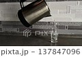 Hot water pouring from metal kettle into clear glass on kitchen counter closeup boiling liquid stream filling transparent cup beverage preparation domestic interior morning routine hydration concept 137847096