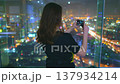 Woman make panoramic shot of night big city lights. Female standing on top floor viewpoint, holding camera photograph the urban skyline. The cityscape features tall buildings, cars, and streets roads 137934214