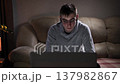 At home in living room teenager sits on a sofa near window, working on his laptop. A night lamp and flowers are on the table next to him. The boy is in a dimly lit room with a window that lets in a 137982867