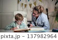 A boy wearing glasses sits in a brightly lit room at a white desk, where his textbooks and notebook are laid out, and works on exercises in his notebook. His mother supervises his studies. She 137982964