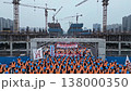 Construction workers in orange vests gather at large urban building site under multiple cranes, holding banners and flags, wearing blue helmets, coordinated salute and rally demonstrating 138000350