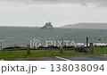 Military frigate sailing on choppy ocean water during an overcast and windy day, with a grassy coastline visible in the foreground and a distant headland on the horizon 138038094