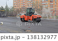 An orange road roller machine compacts fresh hot asphalt on a city street near residential buildings during road construction. 138132977