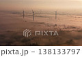 Cinematic aerial view of wind turbines rising above thick morning fog and forest at sunrise. Golden hour light over renewable energy farm. Atmospheric and sustainable future concept video. 138133797