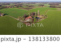 An aerial drone view of a traditional farmhouse with red roofs surrounded by lush green fields. The isolated homestead is nestled among a cluster of trees in a vast, peaceful rural landscape. 138133800