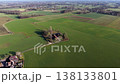 An expansive aerial view showing a secluded farmhouse with red roofing, surrounded by lush green agricultural fields and a nearby village cluster under a clear sky. 138133801