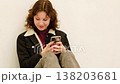 Teenage girl sitting on floor against white wall looking at smartphone screen with serious face, reading messages in chat app and checking social media notifications. 138203681
